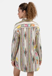 Camicia a righe multicolore con colletto, maniche lunghe e orlo asimmetrico. Presenta motivi intricati in colori e texture vivaci.