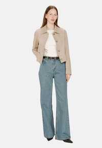 Donna che indossa una giacca beige con bottoni, una maglietta bianca, una cintura nera, dei jeans a gamba larga azzurri chiari e delle scarpe nere a punta, in piedi contro uno sfondo bianco.