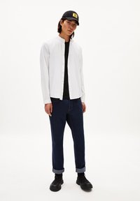 Witte lange mouwen shirt, dichtgeknoopt, over een zwart T-shirt. Blauwe spijkerbroek met opgerolde manchetten en zwarte sneakers. Zwarte pet met logo.