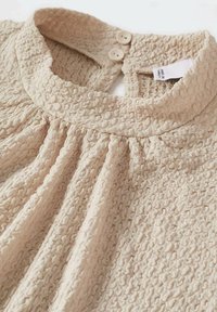 Beige tekstureret top med plisseret design, rund krave og to-knaps lukning bagpå med en "Made in Turkey" etikette.