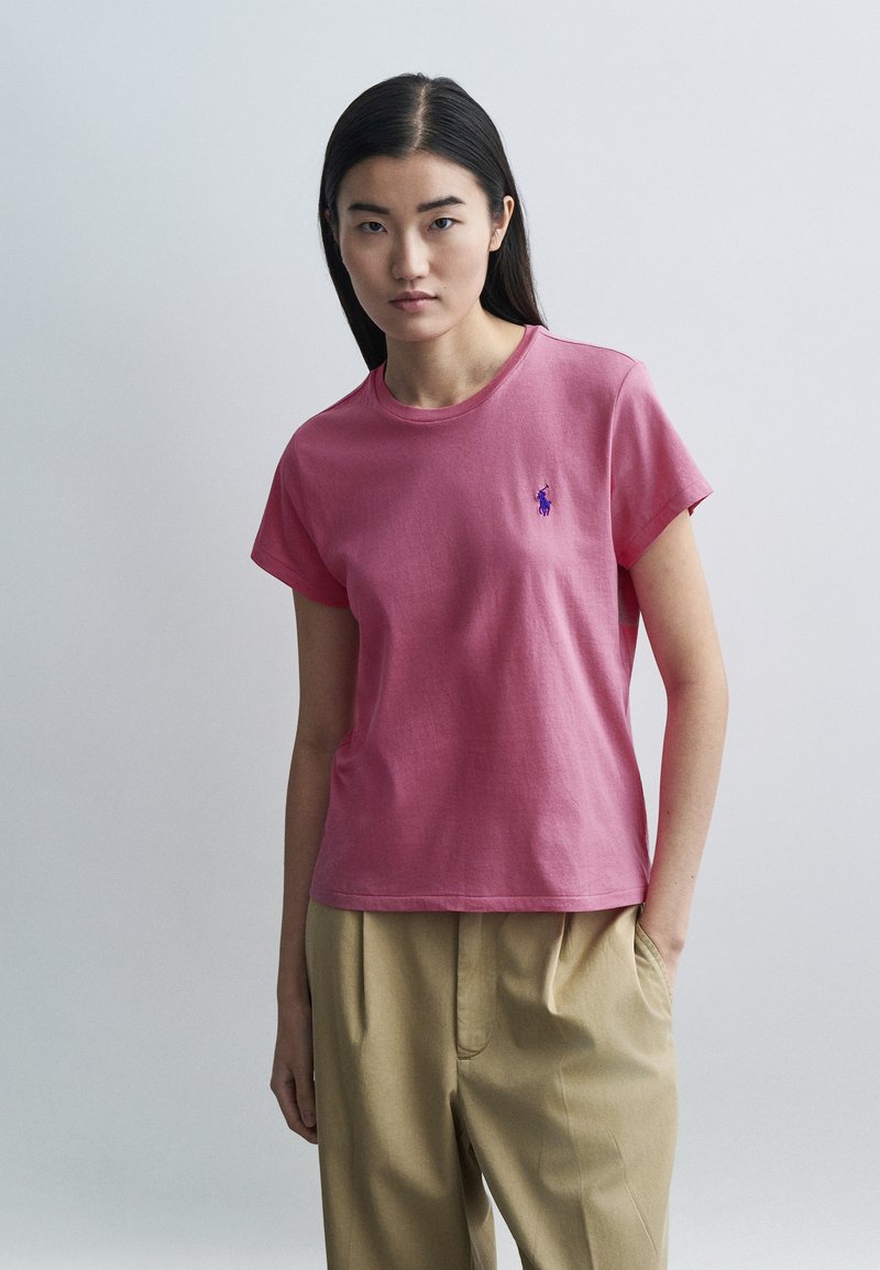 Rózsaszín pamut póló rövid ujjakkal és kerek nyakkivágással, a mellkason egy kis kék hímzett logóval. Bézs nadrággal viselve.