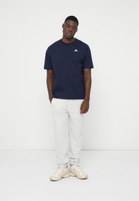 Navyblaues Baumwoll-T-Shirt mit kurzen Ärmeln und weißem Logo, kombiniert mit hellgrauen Sweatpants und beigen Turnschuhen, vor einem schlichten Hintergrund stehend.