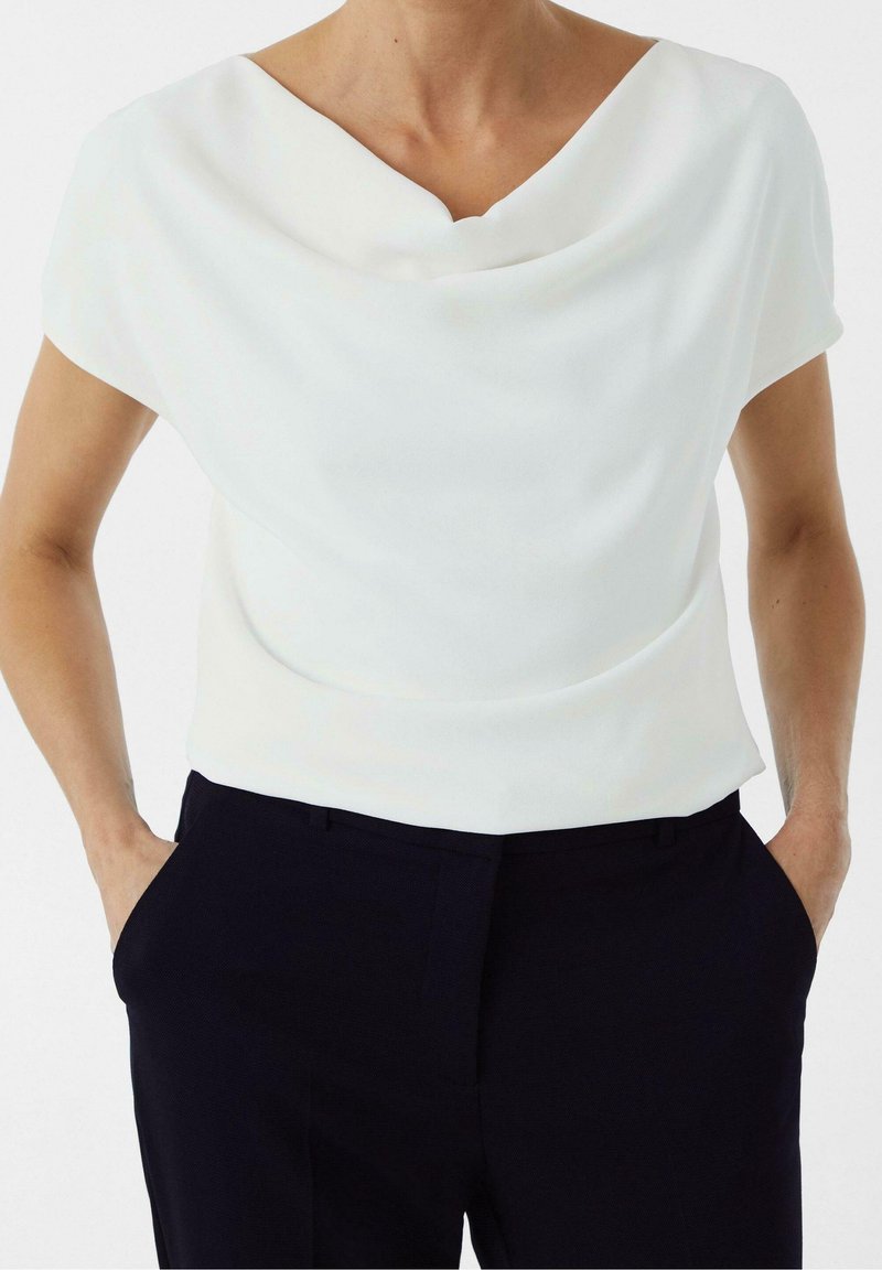 Femme portant un haut blanc à manches courtes col bénitier, rentré dans un pantalon sombre taille haute, les mains dans les poches.
