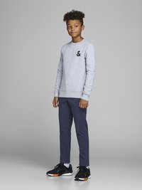 Sudadera gris con logotipo negro, pantalones ajustados azul marino y zapatillas negras con acentos naranjas. Cabello estilizado en rizos cortos.