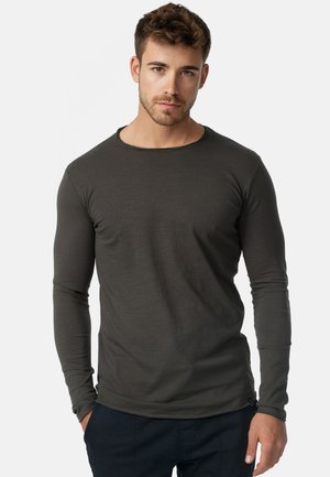 Junger Mann mit kurzen lockigen Haaren, trägt ein eng anliegendes dunkelgraues Langarmshirt und schwarze Hosen, steht vor einem schlichten hellen Hintergrund.