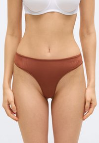 Γυναίκα που φοράει λευκό σουτιέν και καφέ εσώρουχο με ψηλή γραμμή, στέκεται με τα χέρια χαλαρά στο πλάι μπροστά σε απλό φόντο.