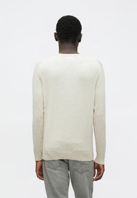 Άνδρας με κοντά σγουρά μαλλιά, φορώντας μια μπεζ μακρυμάνικη μπλούζα και ανοιχτό γκρι τζιν, απεικονίζεται από πίσω σε άσπρο φό�ντο.