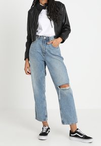 Veste bomber en cuir noir, t-shirt blanc, jeans déchirés larges bleu clair, et baskets noires avec semelles blanches.