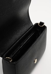 Borsa a tracolla in pelle nera testurizzata con alette aperte, chiusura in ottone dorato e due anelli metallici per il cinturino, che mostra scomparti interni vuoti.
