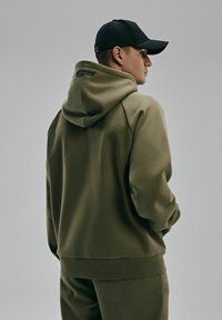 Oliwkowa bluza z kapturem wykonana z miękkiego materiału, z dużym kapturem, ściągaczami przy mankietach i u dołu oraz małym logo z tyłu.