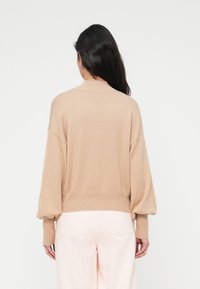 Πλεκτό μπεζ πουλόβερ με άνετη εφαρμογή, χαμηλωμένους ώμους και ριμπ τελείωμα· συνδυασμένο με παντελόνι ανοιχτό ροζ χρώματος· λεία υφή και ελαφρύς σχεδιασμός.