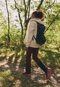Jachetă impermeabilă de culoare deschisă; rucsac închis la culoare; pantaloni vișinii cu panouri negre; încălțăminte de traseu roz; mergând pe o potecă de pământ printre vegetație.
