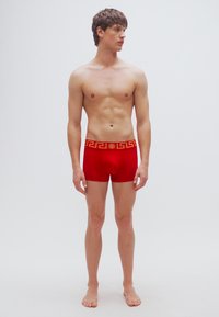 Κόκκινα ανδρικά εσώρουχα μποξεράκια με χρυστό σχέδιο ελληνικού κλειδιού στη μέση. Μαλακό ύφασμα, εφαρμοστή γραμμή και μινιμαλιστικό στυλ. Το μοντέλο στέκεται ξυπόλητο.