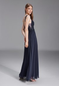 Robe de soirée bleu marine avec corsage en dentelle, dos transparent et jupe fluide. Dispose de fines bretelles et d'un ourlet long au sol. Design élégant.