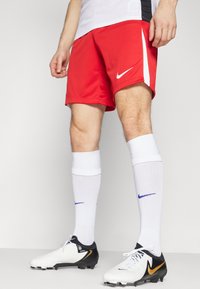Rode sportieve shorts met witte accenten, witte sportsokken en zwart-witte voetbalschoenen met een gouden logo. Gestructureerde stof, aansluitend ontwerp.