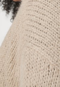 Lichtbeige gebreide stof met een getextureerd ribbelpatroon; zacht, pluizig uiterlijk; subtiele tintvariaties versterken het ontwerp.