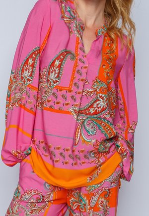 Femme portant un chemisier à motif paisley rose et orange avec un pantalon assorti, montrant des motifs floraux et géométriques.