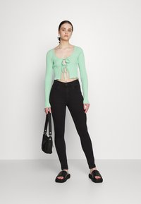 Top cropped canelado verde menta com decote redondo e detalhe em laço na frente, combinado com jeans slim-fit pretos e sandálias slides pretas.
