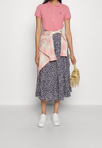 Pink polo shirt, navy floral midi skirt, light plaid wrapped shirt, white sneakers, and a light beige fringe handbag.