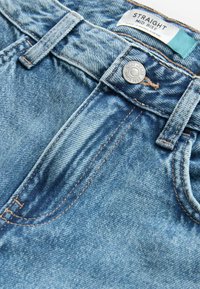 Jeans de mezclilla azul claro con un ajuste de altura media, corte recto, costuras de cobre y cierre de botón. Etiqueta de la marca visible en la parte interior de la cinturilla.