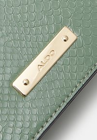 Grön texturerad läderplånbok med en guld rektangulär skylt där "ALDO" är präglat. Sytt kanter och en slät, glänsande yta.