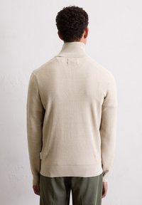 Beżowy sweter dziany z wysokim kołnierzem, prążkowaną strukturą i dopasowanym krojem. Posiada długie rękawy i metkę z boku.