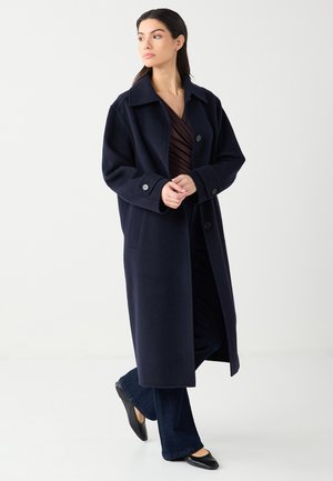 Femme portant un long manteau bleu marine foncé par-dessus un haut sombre et un jean, debout les mains jointes et regardant sur le côté sur un fond uni.