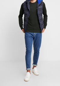 Marineblauer gesteppter Weste mit blauer Kapuze über einem dunkelgrünen Langarmshirt, kombiniert mit hochgerollten blauen Jeans und weißen Turnschuhen.