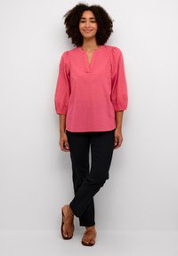Blusa rosa a tre quarti con scollatura a V, caratterizzata da punti a rilievo, abbinata a pantaloni neri e infradito color beige. Sfondo semplice.