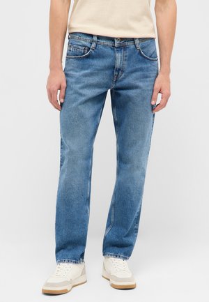 Straight Fit blaue Denim-Jeans getragen mit weißen Sneakers und einem beigen Hemd, gezeigt von der Taille bis zu den Füßen vor neutralem Hintergrund.