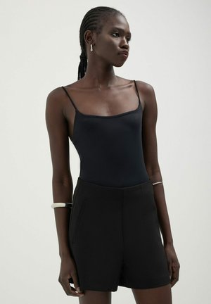 Body nero con spalline sottili, abbinato a pantaloni corti neri dal taglio sartoriale. Texture liscia, design minimale, con tasche laterali inclinate.