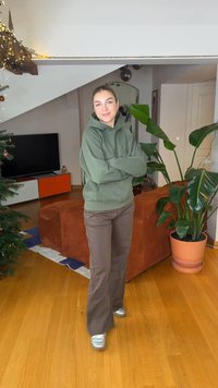 Masivna hoodie maslinasto zelene boje s utisnutim tekstom, smeđe hlače širokih nogavica i srebrne tenisice; unutarnji prostor s vidljivim biljkama i namještajem.