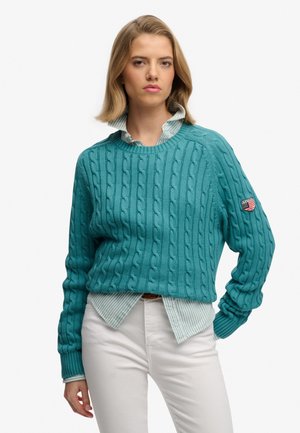 Ung kvinde iført teal kabelstrikket sweater over en stribet skjorte med krave og hvide bukser, stående med den ene hånd i lommen.