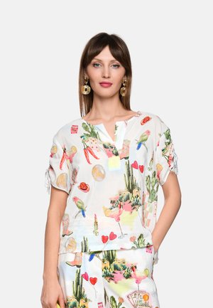 Frau trägt weißen passenden Top und Hose mit bunten Illustrationen von Wüste, Vogel und Cocktail, goldene fächerförmige Ohrringe und glattes braunes Haar.