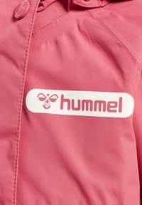 Veste rose avec une texture lisse et mate. Elle présente un logo blanc avec l'inscription "hummel" et une fermeture à boutons ronds.