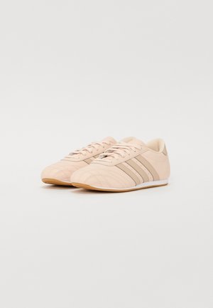 Paire de baskets lacées beige clair avec trois bandes beige plus foncé sur les côtés et semelles couleur gomme sur fond blanc.