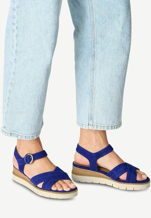 Sandali blu in suede con cinturini incrociati e fibbia alla caviglia. Presentano un tacco a cuneo con una suola di colore chiaro e una suola testurizzata.