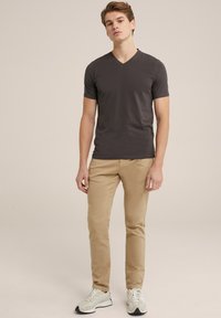 Camiseta de manga corta en gris oscuro con cuello en V, combinada con chinos ajustados beige y zapatillas deportivas de color claro, de pie contra un fondo neutro.