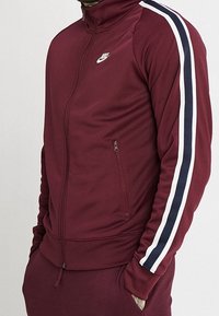 Bordowa rozpinana bluza dresowa z białymi i granatowymi paskami na rękawach. Posiada wysoki kołnierz i zamykaną na zamek kieszeń boczną. Logo Nike na piersi.