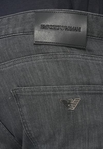 Tmavomodré riflové nohavice s čiernym koženým štítkom na páse, vybaveným vyrazeným nápisom "EMPORIO ARMANI" a strieborným logom na zadnom vrecku.