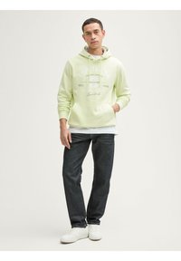 Sudadera verde clara con cordones y bolsillo frontal, que presenta texto y gráficos en blanco. Combinada con jeans oscuros y zapatillas blancas.