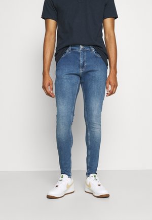 Man staat, draagt blauwe skinny jeans, witte sneakers en een zwart T-shirt met korte mouwen tegen een effen lichte achtergrond.