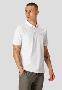 Hvid kortærmet polo shirt i tekstureret stof, med to-knaps knaplukning og et lille sort logo på venstre bryst. Båret med khaki bukser.