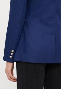 Tummansininen bleiseri, jossa on herringbone-kuviointi, ja kolmella kultaisella napilla hihassa. Yhdistetään mustiin pystysuoriin housuihin.