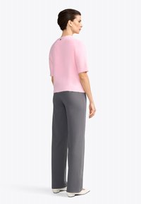 Donna in piedi di lato che indossa un maglione corto rosa a maniche corte, pantaloni grigi a gamba larga, scarpe bianche e orecchini a cerchio dorati su sfondo bianco.