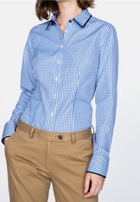 Camisa de cuadros vichy azul y blanca con botones, mangas largas, cuello con solapa y ribete azul marino. Se lleva con pantalones beige, mostrando una textura suave.