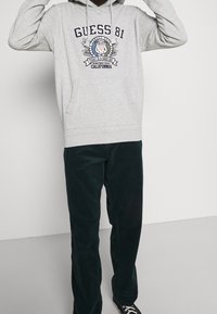 Sudadera gris con capucha, bolsillo frontal y texto impreso, combinada con pantalones de pana acanalada en verde oscuro y zapatillas negras.
