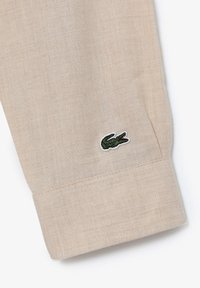 Poignet en tissu beige avec une texture subtile, arborant un logo crocodile vert brodé, des bords soigneusement surpiqués et un drapé souple.