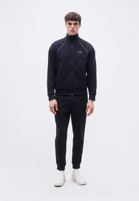 Άνδρας που στέκεται φορώντας μπλε σκούρο μπουφάν με φερμουάρ και λογότυπο BOSS, ταιριαστά μπλε σκούρα παντελόνια φόρμας, λευκές κάλτσες και λευκά αθλητικά παπούτσια, σε απλό φόντο.