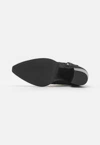 Cizmă de gleznă din suede negru, cu vârful ascuțit, talpă plată și un accent metalic sub formă de cataramă pe lateral. Textură netedă și detalii minime.